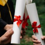 Group of graduates holding diplomas with red ribbons during a ceremony outdoors.